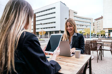 Optimistische Geschäftsfrau, die auf einem Netbook tippt, während sie mit einem anonymen Mitarbeiter am Tisch sitzt und während der Arbeit auf der Terrasse eines Cafés auf einem Tablet surft - ADSF33039