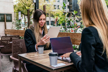 Lächelnde Unternehmerin, die auf einem Tablet surft, während sie mit einem anonymen Kollegen an einem Tisch sitzt und auf einem Laptop tippt, während sie auf der Terrasse eines Cafés miteinander diskutieren - ADSF33041