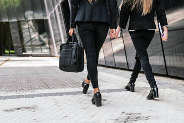 Anonyme weibliche Mitarbeiter mit Tasche und Notizblock in den Händen in stilvoller Kleidung zu Fuß auf gepflasterten Straße entlang modernen Gebäude in der Stadt - ADSF33042