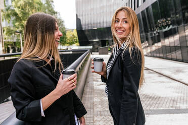 Side view of positive female coworkers with takeaway hot beverages while standing on street near building - ADSF33043