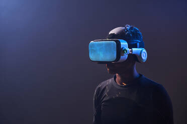 Erstaunte afroamerikanische Spielerin mit moderner VR-Brille vor dunklem Hintergrund im Studio, während sie die virtuelle Realität in Neonbeleuchtung erlebt - ADSF33045