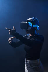 Afroamerikanische Frau mit modernem VR-Headset vor schwarzem Hintergrund mit erhobenen Händen beim Erkunden der virtuellen Realität im Studio - ADSF33047