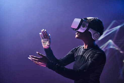 Afroamerikanische Frau mit modernem VR-Headset vor schwarzem Hintergrund mit erhobenen Händen beim Erkunden der virtuellen Realität im Studio - ADSF33048