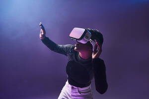 Erstaunte afroamerikanische Spielerin mit moderner VR-Brille vor dunklem Hintergrund im Studio, während sie die virtuelle Realität in Neonbeleuchtung erlebt - ADSF33050
