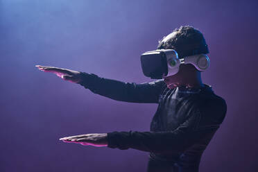 Erstaunte afroamerikanische Spielerin mit moderner VR-Brille vor dunklem Hintergrund im Studio, während sie die virtuelle Realität in Neonbeleuchtung erlebt - ADSF33051
