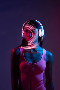 Charmante brasilianische Frau, die mit drahtlosen Kopfhörern Musik hört, während sie stehend in einem obskuren Studio auf dunklem Hintergrund mit Lichtschatten auf dem Gesicht wegschaut - ADSF33058