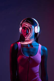 Charming Brazilian female listening to music in wireless headphones while standing looking away in obscure studio on dark background with shadow of light on face - ADSF33058