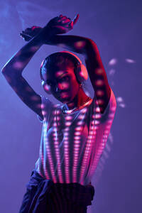 Charming Brazilian female listening to music in wireless headphones while dancing on purple background in studio with shadows on disco ball and fluorescent lights - ADSF33059