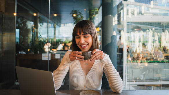Junge positive Frau in lässigem Outfit sitzt am Tisch in einem Café mit einer Tasse Kaffee in der Hand und schaut auf den Bildschirm eines Laptops während eines Videoanrufs - ADSF33074
