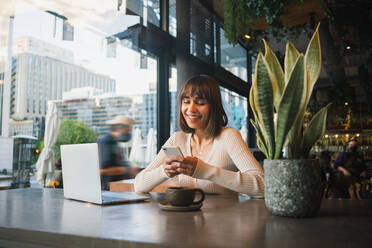 Junge positive Frau in legerem Outfit sitzt am Tisch in einem Café mit einer Tasse Kaffee und schaut auf den Bildschirm eines Laptops, während sie auf einem Smartphone surft - ADSF33077