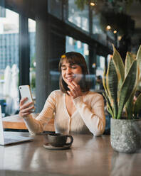 Junge positive Frau in legerem Outfit sitzt am Tisch in einem Café mit einer Tasse Kaffee und schaut auf den Bildschirm eines Laptops, während sie auf einem Smartphone surft - ADSF33078