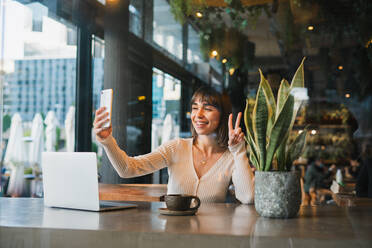 Junge lächelnde Frau in Freizeitkleidung, die ein Selfie-Foto auf ihrem Smartphone mit einer Siegesgeste der Finger macht, während sie mit einer Tasse Kaffee und einem Laptop am Tisch sitzt - ADSF33079