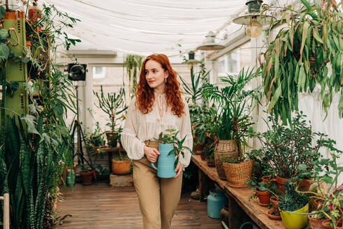Charming female with long ginger hair standing with jug of seedling in indoor garden with various potted houseplants at home - ADSF33080
