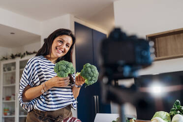 Smiling vlogger showing broccoli filming through camera at home - MEUF05083