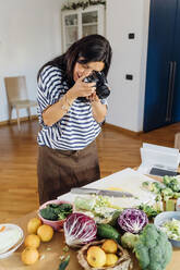 Frau fotografiert frisches Gemüse mit Kamera zu Hause - MEUF05089