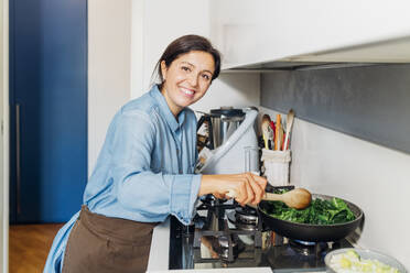 Smiling woman cooking spinach in frying pan on stove at home - MEUF05110