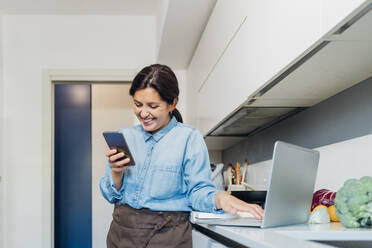 Lächelnde Frau mit Mobiltelefon und Laptop an der Küchentheke zu Hause - MEUF05112