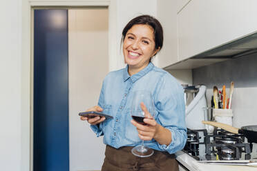 Happy woman with smart phone holding wine glass in kitchen at home - MEUF05114