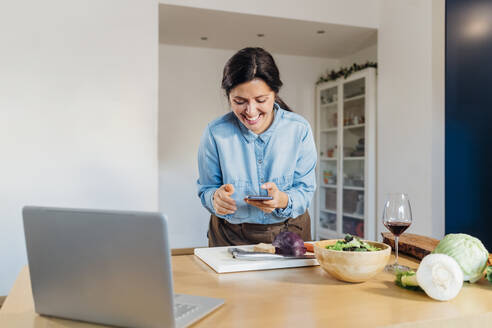 Smiling woman using smart phone standing at table - MEUF05120