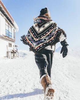 Man wearing poncho running on snow towards mountain hut - OMIF00334