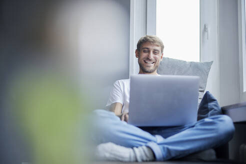 Smiling businessman using laptop sitting on sofa at home - FMKF07364