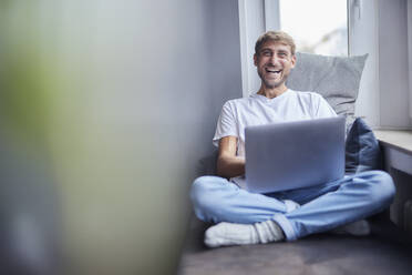 Lachender Geschäftsmann mit Laptop auf dem Sofa zu Hause - FMKF07365