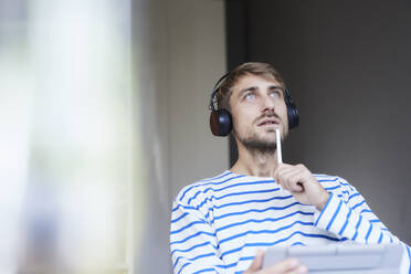 Thoughtful businessman with headphones holding tablet PC at home office - FMKF07372