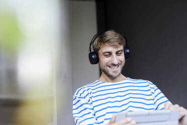 Lächelnder Geschäftsmann mit drahtlosen Kopfhörern, der im Büro zu Hause einen Tablet-PC benutzt - FMKF07373