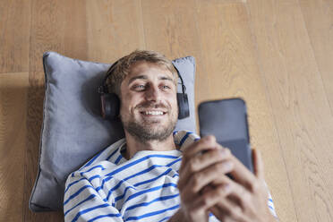 Lächelnder Mann mit Kopfhörern und Smartphone auf Hartholzboden liegend - FMKF07377