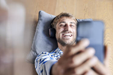 Glücklicher Mann mit Kopfhörern, der ein Selfie mit seinem Smartphone zu Hause macht - FMKF07379