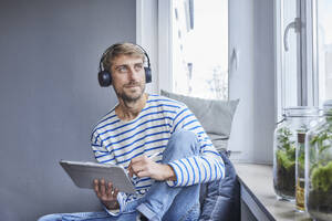 Kontemplativer Geschäftsmann mit Tablet-PC, der zu Hause am Fenster sitzt - FMKF07381