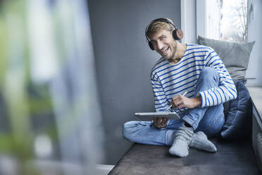 Happy businessman with tablet PC sitting on sofa at home - FMKF07385