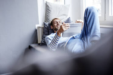 Smiling businessman using tablet PC lying on sofa at home - FMKF07386