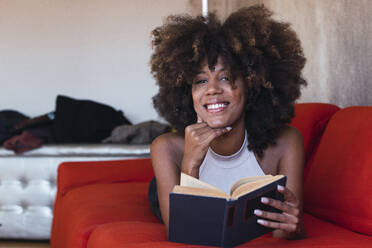 Smiling woman with book lying on sofa - PNAF02655