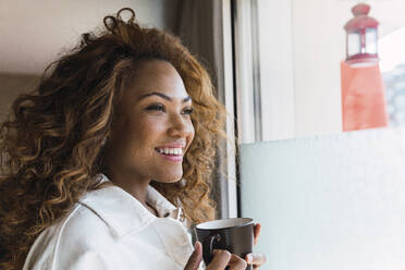 Young smiling woman holding coffee cup at home window - PNAF02675