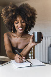 Junge Frau mit Kaffeetasse, die zu Hause in ein Buch schreibt - PNAF02680