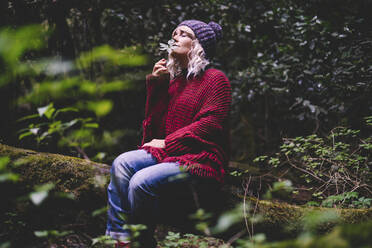 Frau, die an einem Blatt riecht, sitzt auf einem Baumstamm im Wald - SIPF02703