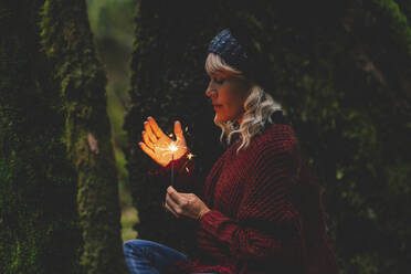 Woman wearing warm clothing holding sparkler in forest - SIPF02709