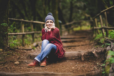 Smiling woman with hands on chin sitting in forest - SIPF02714