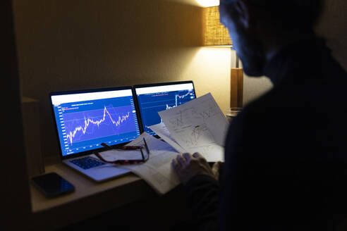 Businessman with documents analyzing stock market data on laptop at home office - JPTF00995