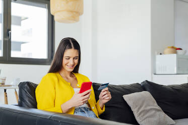 Smiling woman doing online shopping on smart phone in living room - PGF00932