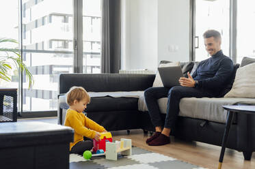 Father freelancing on laptop and son playing in living room - PGF00940