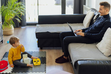 Ein kleiner Junge sieht seinen Vater an, der im Wohnzimmer an einem Laptop arbeitet - PGF00941