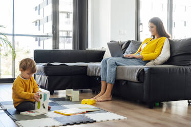 Mutter arbeitet am Laptop, während ihr kleiner Junge zu Hause spielt - PGF00946