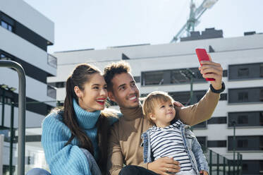 Glückliche Familie, die ein Selfie mit ihrem Smartphone vor einem Gebäude macht - PGF00965