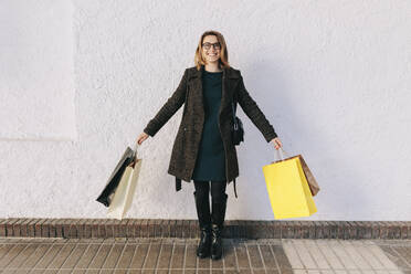 Lächelnde Frau steht vor einer weißen Wand und hält Einkaufstüten - MRRF01829