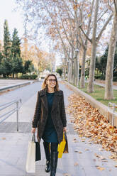 Lächelnde Frau mit Einkaufstüten auf dem Fußweg im Herbst - MRRF01830
