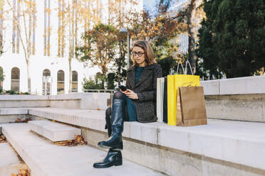 Woman using smart phone sitting on steps with shopping bags - MRRF01831