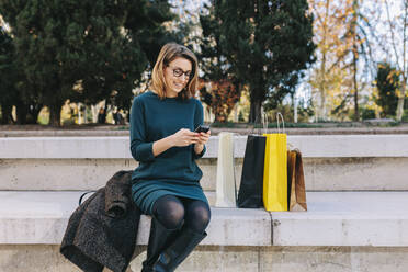 Lächelnde Frau, die ein Smartphone benutzt und auf einer Treppe neben Einkaufstüten sitzt - MRRF01832