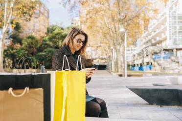 Woman using smart phone sitting by shopping bags - MRRF01835